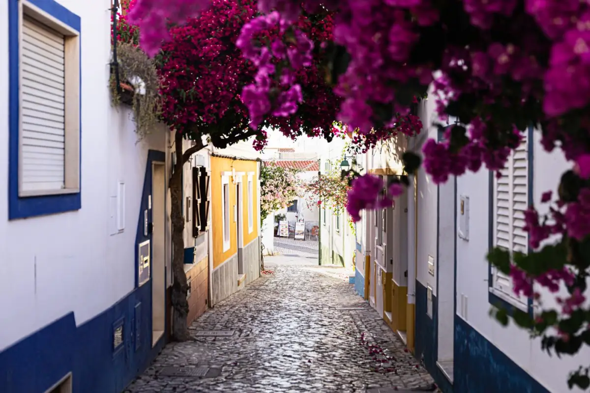 Rua estreita com buganvílias roxas entre casas brancas de Ferragudo, vila de pescadores do Algarve