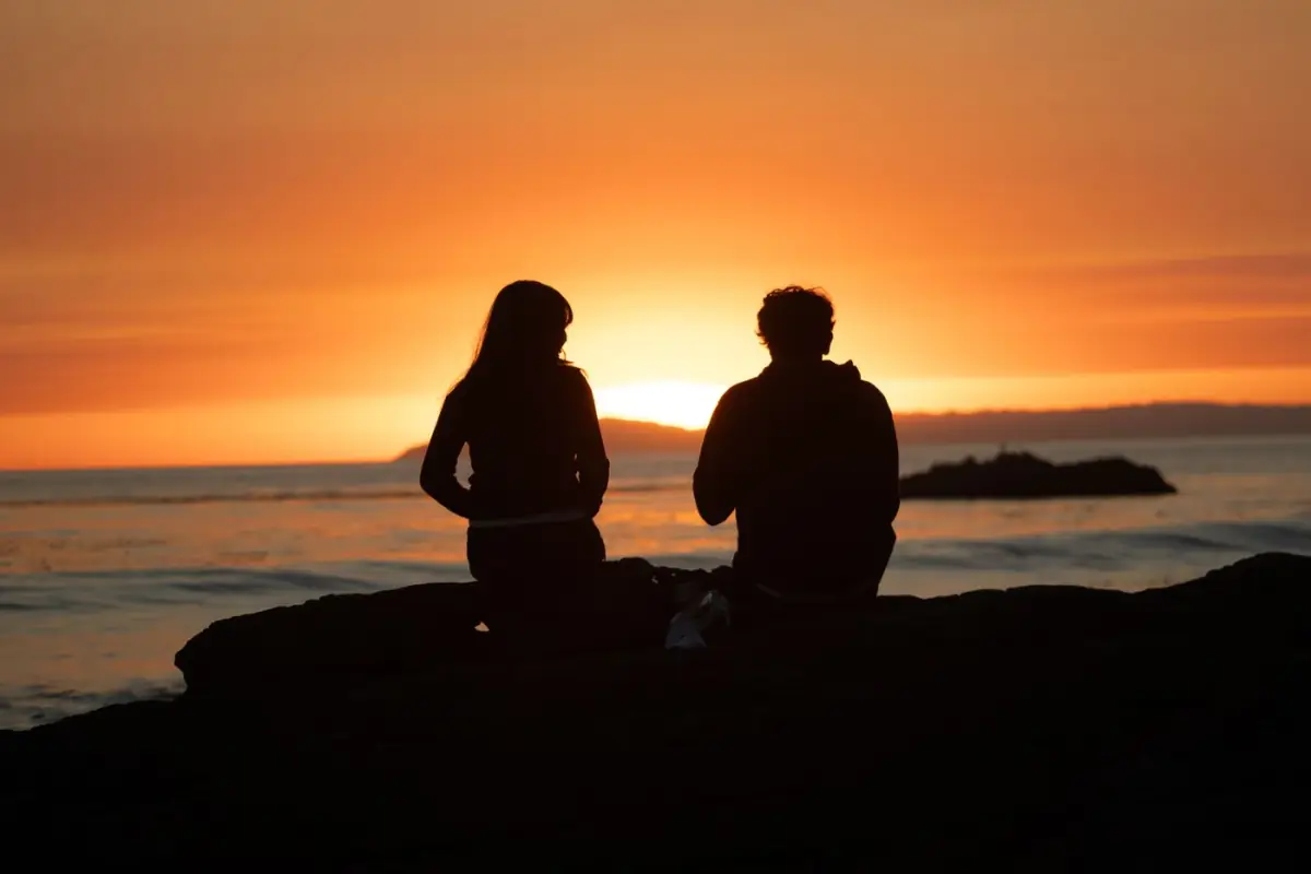 Casal sentado numa rocha de uma falésia algarvia a ver o pôr do sol sobre o mar