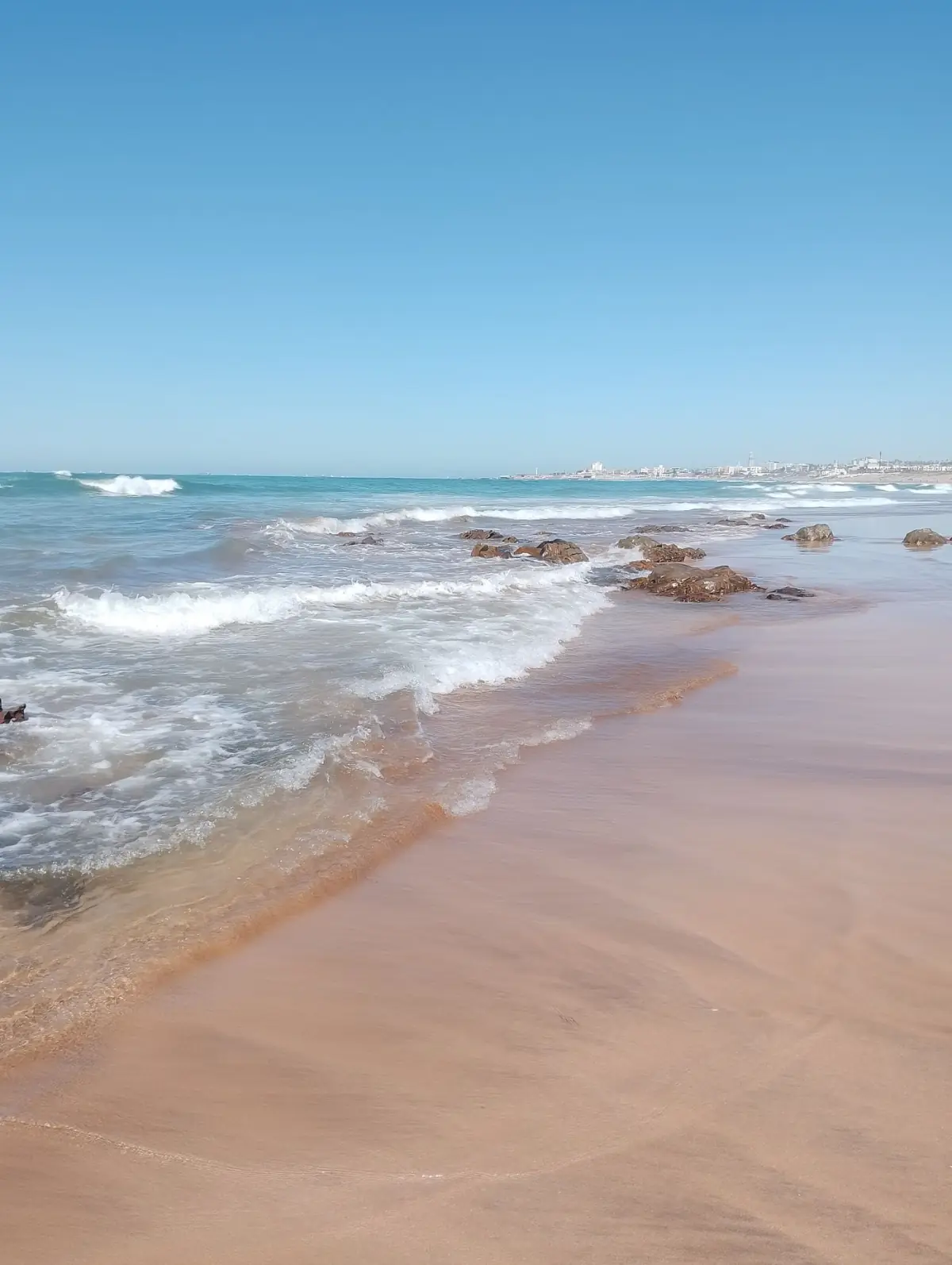 Areal largo e sereno da Praia do Barril com linha de rochas baixas e mar calmo do Sotavento, em Tavira